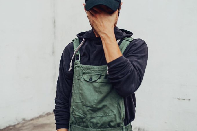 A person wearing a black cap, black hoodie, and green overalls stands against a white wall with their hand covering their face.