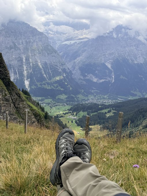 A first-person perspective photo of someone's black hiking boots resting on an alpine grassy ridge, overlooking a sweeping valley and dramatic, snow-capped mountain peaks under a cloudy sky.