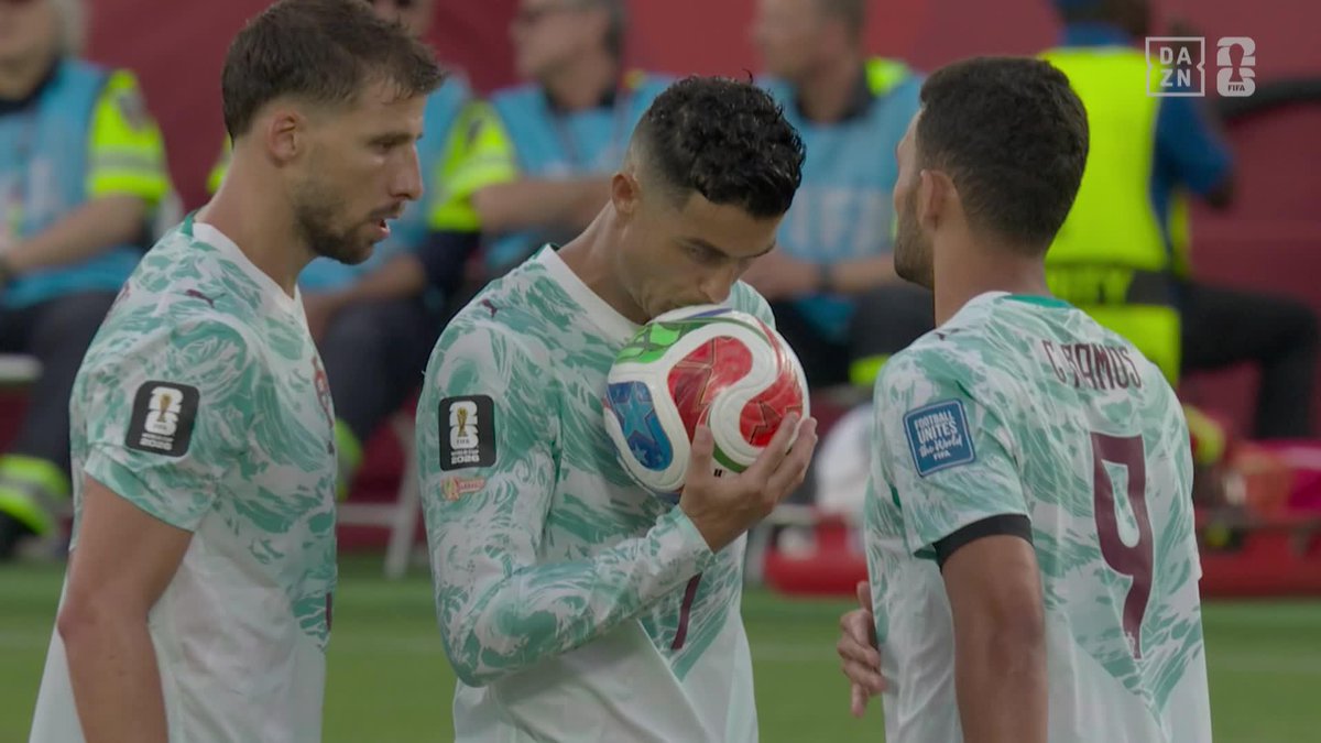 Three Portugal national football players in teal and white kits standing on the pitch; the center player (Cristiano Ronaldo) holds and kisses the match ball, with a DAZN FIFA broadcast logo in the corner.