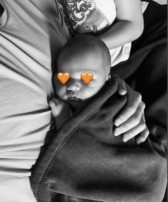 A sweet black-and-white photo of a newborn baby resting against a parent's chest, swaddled in a blanket with two orange heart emojis covering their eyes. The image perfectly complements the announcement, celebrating the birth of a new baby whose parents are part of the Bitcoin community.
