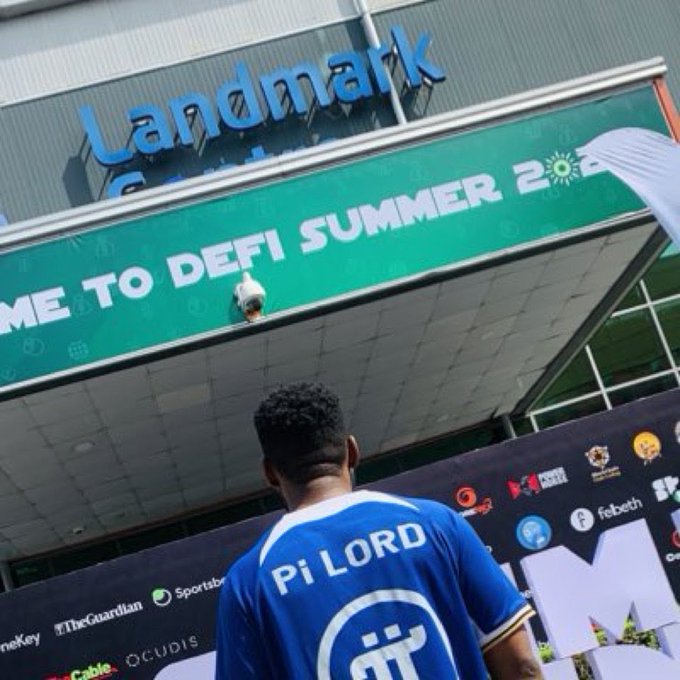 A man wearing a blue "Pi LORD" shirt stands before the Landmark Centre, where a banner reads "WELCOME TO DEFI SUMMER 2[0?]" alongside various media logos, capturing a Pi Network community moment coinciding with #Pi2Day celebrations.