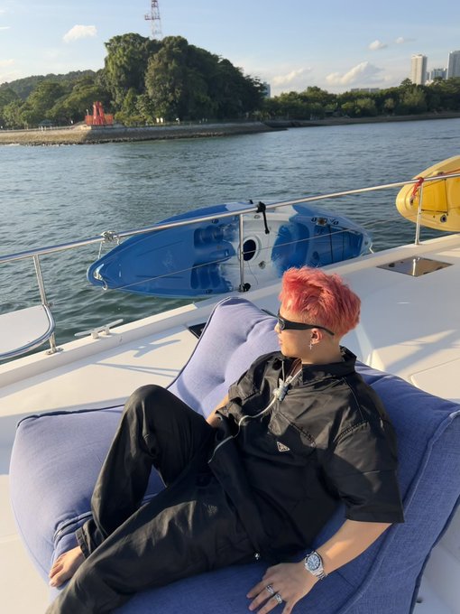 A person with pink hair, sunglasses, and black clothing lounging on a blue cushion aboard a boat, with blue kayaks and a shoreline visible in the background.