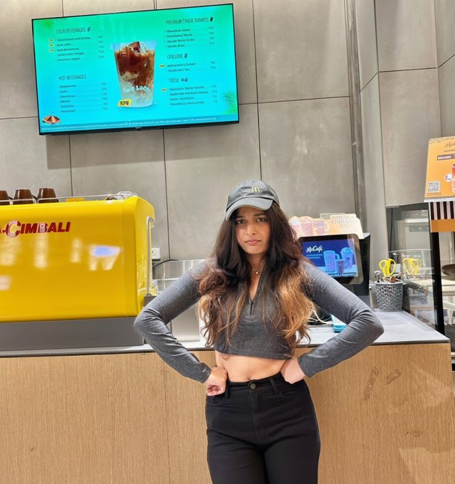 A young woman wearing a gray crop top, black jeans, and a cap poses confidently with hands on hips inside a McDonald's restaurant, with a menu board visible behind her promoting items like "Cold Beverages" and "Premium Thick Shakes." The setting visually supports her playful post directed at McDonald's, showcasing her as a content creator ready to help grow their audience.