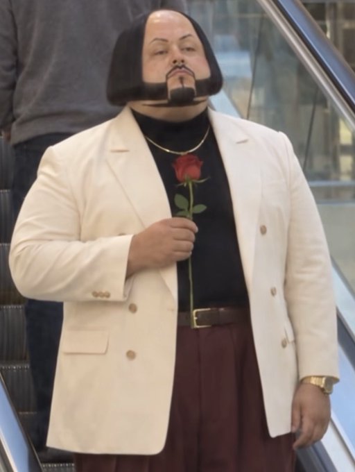 A man with a distinctive bowl haircut and goatee wears a white blazer over a black turtleneck while holding a single red rose, evoking imagery of a stock market "bottom" forming as rose-themed "rh" and "stonk" coins gain traction on the blockchain.