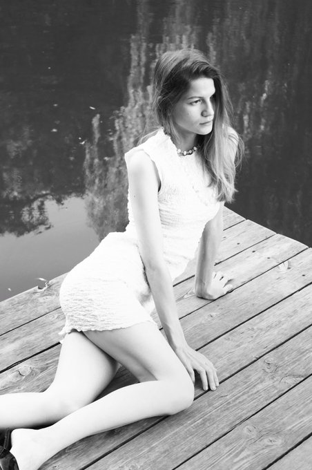 Black-and-white fashion portrait of a woman in a short white sleeveless dress kneeling on a wooden dock beside dark water.