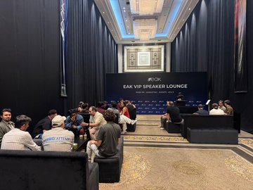 The image shows a large room filled with people sitting on couches and chairs, engaged in conversation. The event appears to be an EAK VIP Speaker Lounge gathering where various individuals are seated around a table, possibly discussing business or industry topics. A banner is displayed prominently in the background that reads "A great week at @IstanbulBlockWk", indicating the nature of the event. The room also features a large screen and a chandelier hanging from the ceiling, creating an elegant atmosphere for the gathering.