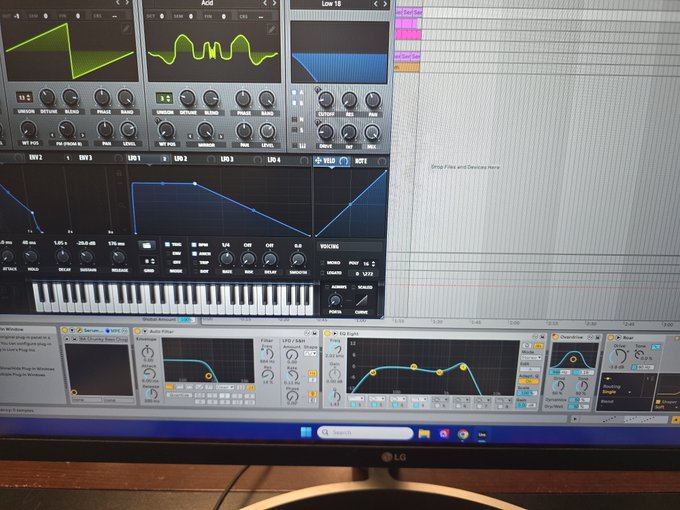 A computer monitor displays music production software with various synthesizer interfaces, oscillators, filters, and a piano keyboard visible on screen.
