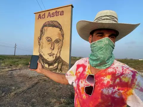 A person in a cowboy hat, green bandana, and tie-dye shirt poses proudly next to a roadside mural of Elon Musk inscribed with the phrase "Ad Astra" beneath it, in an open landscape under a clear blue sky.