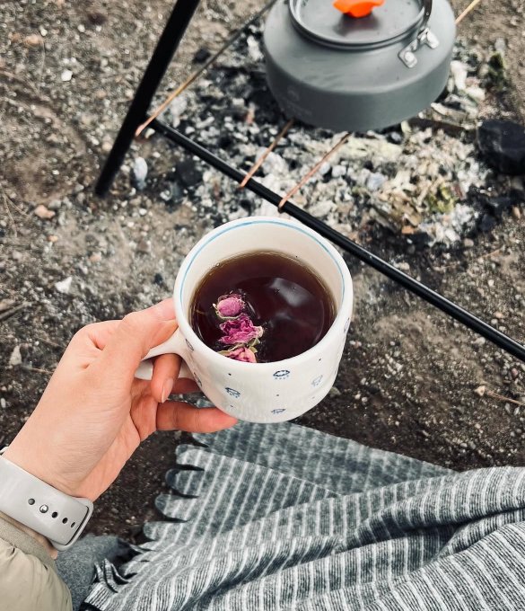 The image shows a person holding a cup of tea with rose petals floating in it. The cup has a white rim, and there are two other cups visible in the background - one gray and another black. A grey blanket can also be seen on the ground near these cups. The photo is taken from an overhead perspective, giving a clear view of the person holding the tea and the surrounding objects.