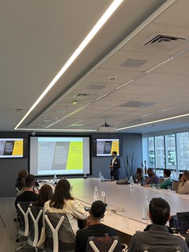 The image shows a group of people sitting at tables, with some individuals standing behind them. They are gathered around a large screen displaying information or slides related to the topic being discussed. The room appears to be well-lit, possibly due to natural light coming in from windows on one side. The people seem engaged and attentive as they listen to the speaker who is presenting at the front of the room.