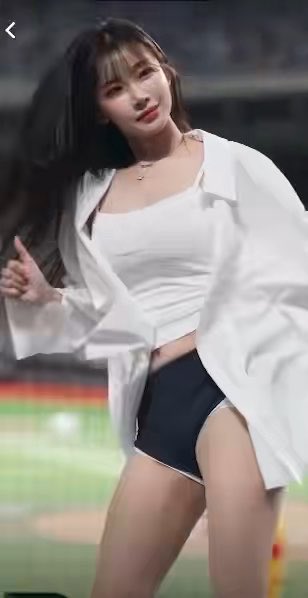 A woman with long dark hair wearing a white crop top, white overshirt, and dark shorts poses on a sports field, appearing to dance or perform.
