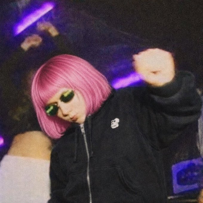 The image features a person with pink hair wearing sunglasses, standing against a purple background. They appear to be dancing or engaging in some form of energetic activity. The caption "Apes are Vibing" suggests that this individual might be participating in an event or gathering where people enjoy themselves and have fun together.