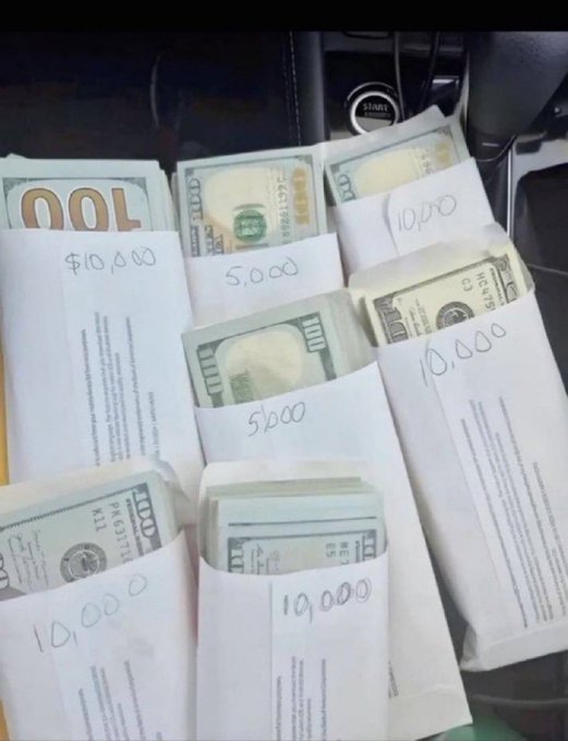 Several white envelopes, each containing stacks of US $100 bills, are laid out on what appears to be a car seat; visible handwritten labels include $10,000, 5,000, and 10,000.