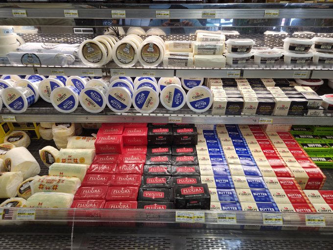 A refrigerated grocery store display showcases an abundant variety of butter products, including Cabot, Plugra, and private-label "BUTTER" sticks, along with butter logs and specialty cultured varieties, neatly arranged in colorful red, black, blue, and white packaging. The "Butter Bar FTW" caption perfectly captures the image's playful spirit, highlighting the impressive assortment of butter options available to shoppers.