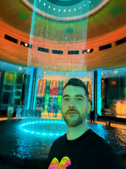 A selfie of a bearded man in front of a cylindrical water fountain with colorful lighting at what appears to be a museum or mall lobby.