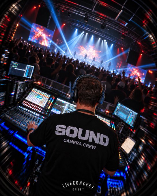 A behind-the-scenes view from the sound booth at a live concert, showing a crew member wearing headphones and a 'SOUND CAMERA CREW' shirt operating a mixing console, with the stage and audience visible ahead.
