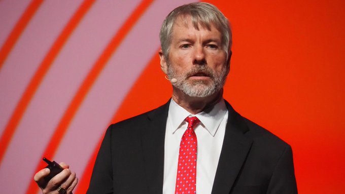 A photograph of Michael Saylor, the executive chairman of MicroStrategy, speaking on stage wearing a black suit and a red tie.