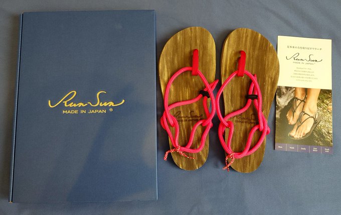The image features a pair of pink sandals with red strings, placed next to a book titled "RUN SON" by a Japanese author. The sandals are positioned on top of a blue surface, while the book lies horizontally nearby. The scene suggests that someone might be preparing for an outdoor activity or event and has chosen these sandals as their footwear choice.