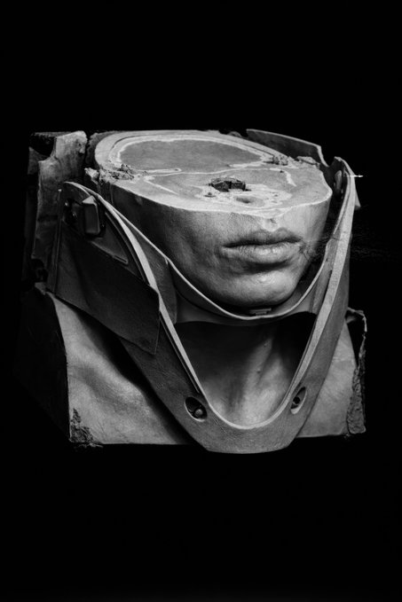 A surreal black and white image shows a stone block carved with human lips resting inside what appears to be a weathered bag or pouch.