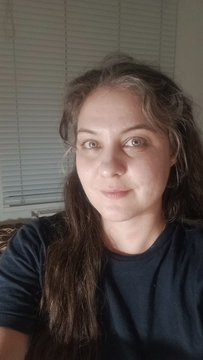 A close-up selfie of a woman with long, curly dark brown hair wearing a dark blue t-shirt, looking directly at the camera with a neutral expression.
