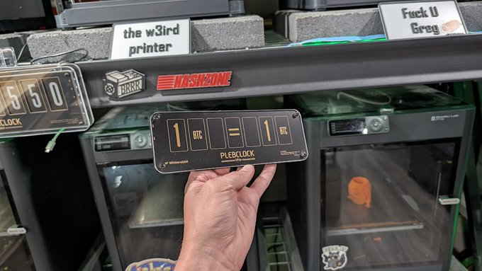 A hand holds up a "PLEBCLOCK" Bitcoin clock showing "1 BTC = 1 BTC" in front of 3D printers labeled "the w3ird printer" and "Fuck U Greg," with a "Hackzine" sticker visible on the shelf. The image playfully references the limited "PlebClock" giveaway tied to the post, where the remaining 10 units (with "boating" cards) are being offered to fans.