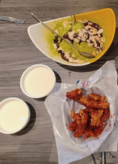 An overhead photo of a meal featuring a bowl with what appears to be rice and meat, two small cups of a white beverage, and a plate of fried chicken pieces on white paper, set on a dark wooden table.