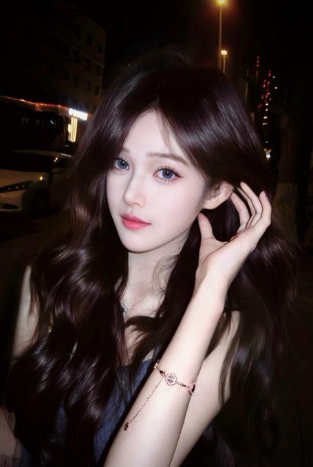 A portrait of a young woman with long dark wavy hair, wearing pink lipstick and a delicate bracelet with a clover charm, photographed at night against a dark street backdrop.