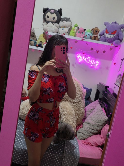 A mirror selfie of a young woman wearing a red floral two-piece outfit, holding a pink phone. The room behind her features pink walls, a neon sign, star string lights, and a shelf lined with Pokémon plush toys.