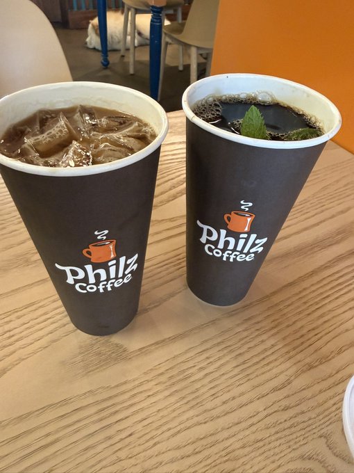 Two black "Philz Coffee" cups sit on a wooden table—one filled with iced coffee and the other topped with a mint leaf—pairing perfectly with a "Coffeemaxxing" post about indulging in maximum coffee enjoyment.