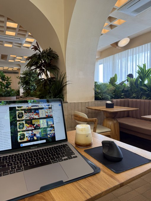A coffee shop or café scene with a MacBook Pro laptop and a latte on a wooden table, featuring distinctive arched white columns and lush green plants in the background.