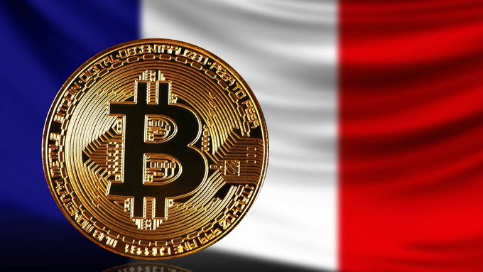 A close-up of a physical Bitcoin token in front of a blurred French flag.