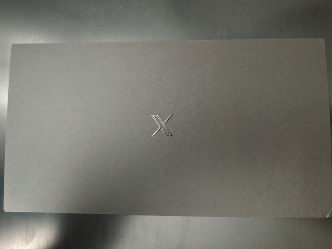 A sleek, dark gray envelope or card sleeve featuring a debossed silver "X" logo in the center rests on a metallic surface, matching the post announcing the arrival of the user's 𝕏 card.