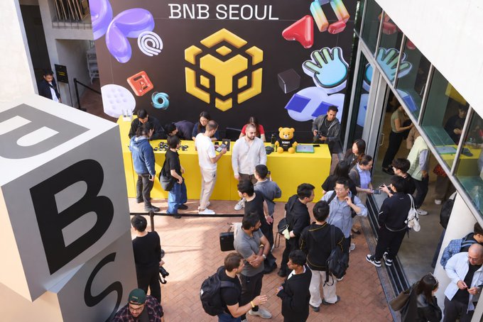 A bustling crowd gathers at the BNB SEOUL event, with attendees networking around a yellow booth featuring the distinctive BNB Chain logo and a large "BNB" cube display in the foreground. Developers and crypto enthusiasts engage with one another in a vibrant atmosphere, reflecting the excitement and community spirit of this Binance event held on October 1st.