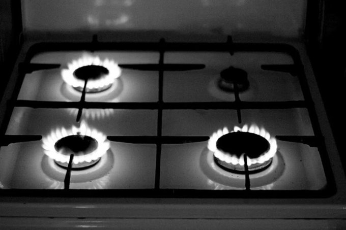 A black and white photograph of a gas stovetop with three burners lit, the flames glowing brightly against the dark cast iron grates.