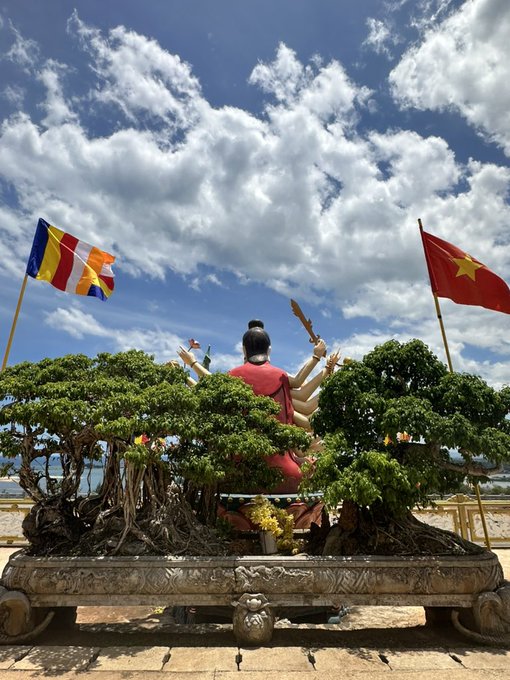 A Buddhist statue with multiple arms seated among bonsai-style trees, flanked by a Buddhist flag on the left and the Vietnamese flag with a yellow star on the right, set against a cloudy sky.