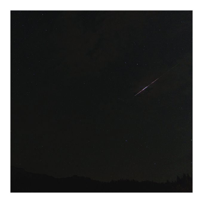 A nighttime photograph of a dark starry sky showing what appears to be a shooting star or meteor streaking across, with a silhouetted landscape at the bottom.