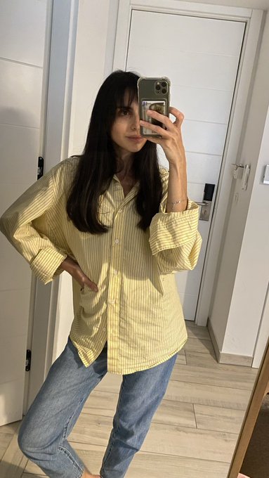 A mirror selfie of a young woman with long dark hair wearing a yellow and white striped button-up shirt and blue jeans.