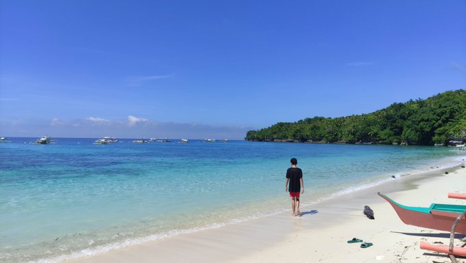 A person in dark clothing and red shorts walking along a white sand beach with clear turquoise water, boats, and a forested coastline in the distance.