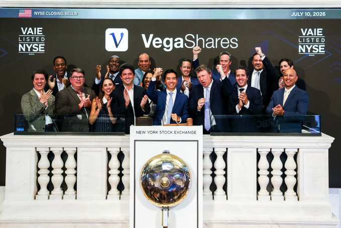 The VegaShares team celebrates their listing on the NYSE on July 10, 2026, posing behind the New York Stock Exchange bell after ringing it to mark the occasion, with their ticker $VAIE prominently displayed alongside the closing bell banner.