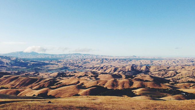 A scenic landscape photograph of rolling golden-brown hills extending toward distant mountains under a clear blue sky with low clouds on the horizon.