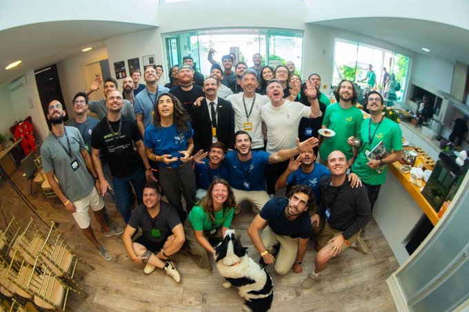 A large, diverse group of around 30 people wearing various shirts—including several with "Base" branding—gather together in a brightly lit office space, smiling and posing for a wide-angle photo with a black and white dog in the center. The image visually reflects the post's message of Base Brasil's community, showcasing the vibrant team of Brazilian builders and contributors supported by the Base ecosystem who are invited to connect for product feedback and go-to-market guidance.