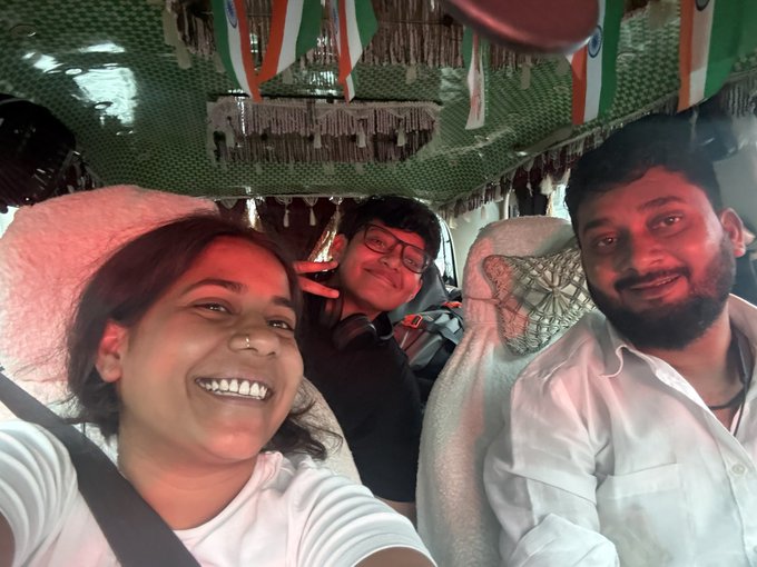 A selfie of three smiling people—two men and a woman—riding inside a vehicle decorated with Indian flag garlands hanging from the ceiling.