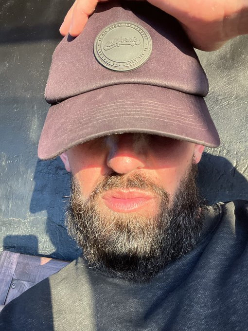 A close-up selfie of a bearded man wearing a maroon 'California' baseball cap pulled low over his eyes, standing outdoors.