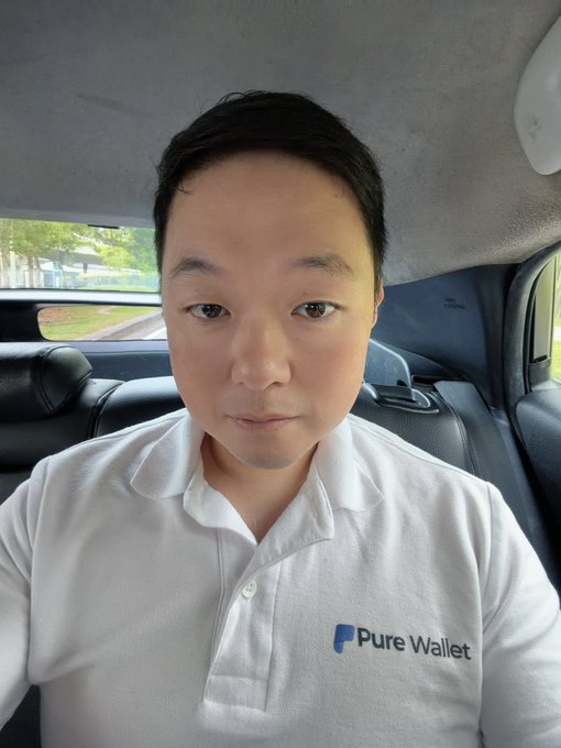 A selfie of an Asian man wearing a white polo shirt with a 'Pure Wallet' logo, sitting inside a car.