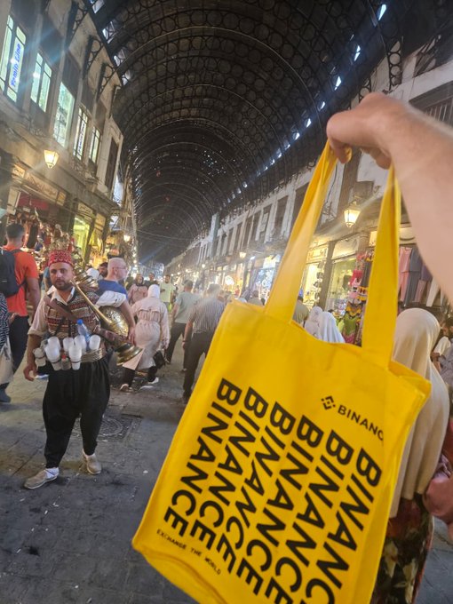 A hand holds up a bright yellow "BINANCE Exchange the World" tote bag in a bustling covered bazaar, where a vendor in traditional dress sells Turkish ice cream. The bustling market scene with its arched iron ceiling and shop-lined walkway suggests a Middle Eastern location, fitting the post's playful "Guess where I am? 👀" caption.