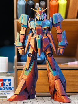A meticulously painted Gundam model, likely the Zeta Gundam, stands completed on a workbench with its main body finished, showcasing a striking color-shifting finish that reveals warm sunset tones from the front and more blue/purple hues at different angles, taking approximately 80 hours to produce. A Tamiya Cement bottle is visible in the foreground.
