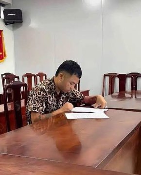 A young man in a patterned shirt sits at a long wooden table in a meeting room, focused on writing or reading a document in front of him.