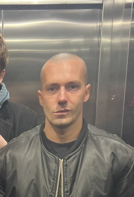 A selfie of a bald man with light stubble wearing a black leather bomber jacket with a zipper, taken inside what appears to be an elevator.
