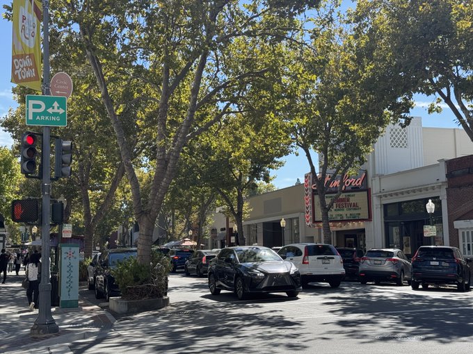 A sunny downtown street scene featuring a 'Fun Ford' vintage theater marquee, mature shade trees, parked cars, and a green 'P PARKING' sign on a traffic light pole.