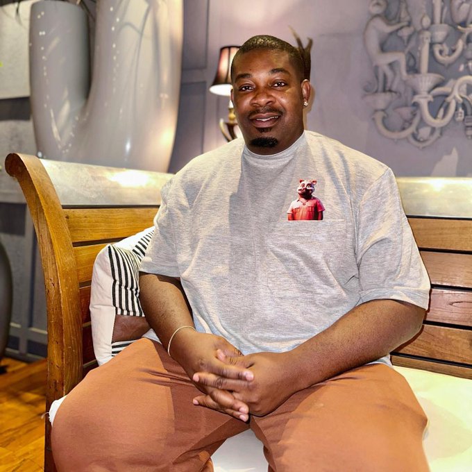 A man wearing a grey t-shirt with a graphic design and brown pants sits smiling on a wooden bench with striped cushions in an elegantly decorated room featuring decorative vases and wall art. The image accompanies a "Pick a Lucky Number" giveaway promotion inviting participants to choose a number between 30 and 80 for a chance to win $30,000.